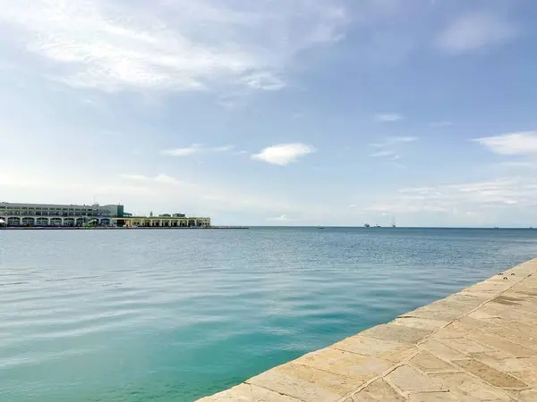 Adriyatik kıyılarının sakin su yüzeyinin panoraması, Trieste şehrinin gezinti güvertesine yakın, ancak bulutlu mavi bir gökyüzünün arka planına karşı..