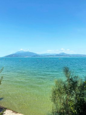 Deniz ufkunun kıyı ağaçlarının arasından göl ve güneşli mavi gökyüzünü birbirine bağlayan bir manzara..