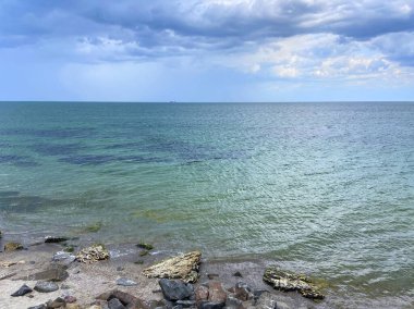 Tehlikeli bir kayalık sahilin manzarası ufuktaki gri fırtına öncesi bulutların arka planında şeffaf deniz mavisi ile hafifçe yıkanır..