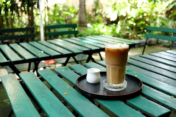 A glass of ice coffee with syrup in white cup on black round plate at green painted wood table in the garden.