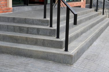 Kare zemin katın etrafındaki gri merdivenler ve binayla beton patika arasında siyah metal korkuluklar boyanmış.,