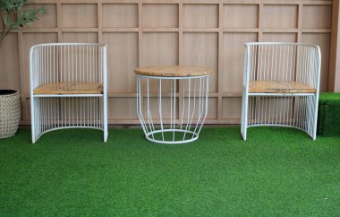 Sandalyeler ve yuvarlak masa yeşil suni çim zemin üzerinde açık kahverengi ahşap zemin. Metal ve ahşaptan yapılmış sandalye ve masa.