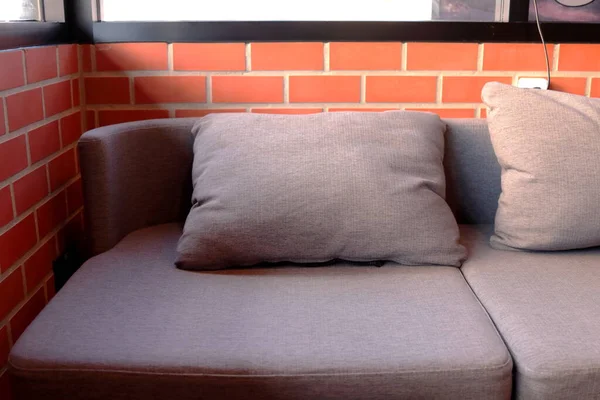 Brown pillow on brown sofa in corner and orange brick wall with sunlight from outside through glass on sofa.
