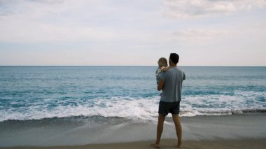 Baba çocuğu kollarına alır ve uzaktaki denize bakar sırtı kameraya dönük bir şekilde. Yüksek kalite 4k görüntü
