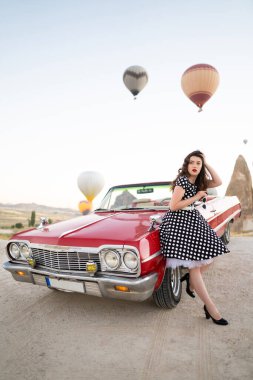 beautiful girl in retro style posing near a vintage red cabriolet car on background of balloons in Cappadocia