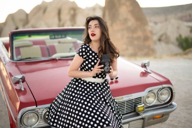 beautiful girl in retro style posing near vintage red cabriolet car with old film camera in hands