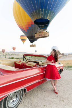 beautiful girl in retro style posing near a vintage red cabriolet car on background of balloons in Cappadocia