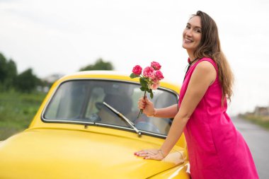 Pembe elbiseli güzel bir kız, sarı retro bir arabanın yanında kırmızı güller açıyor.