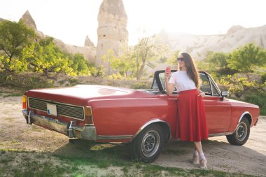 Kapadokya dağlarında eski arabanın yanında içki kutusuyla poz veren etekli güzel mutlu kız.