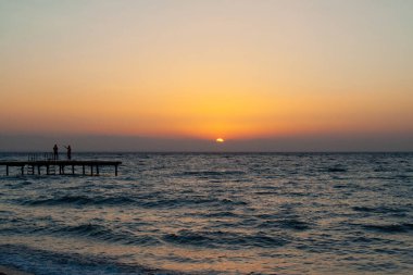 Gün batımında sahil iskelesinde yürüyen insanların silueti.