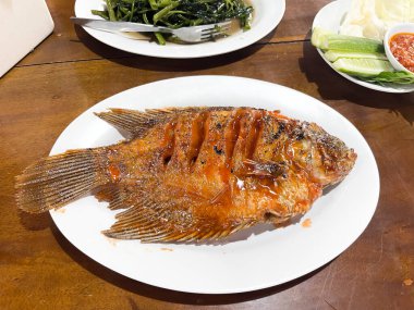 Izgara gourami veya Gurame bakar kırmızı barbekü sosu, sebze ve biber sosu ile masada yedili beyaz bir tabakta.