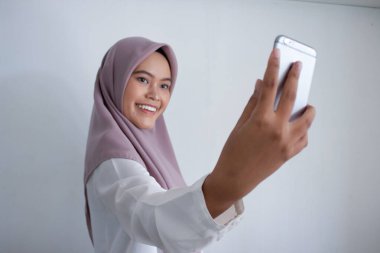 Mor başörtüsü takan Asyalı genç bir kadın telefonuyla selfie çekiyor. Gülümsüyor ve mutlu.
