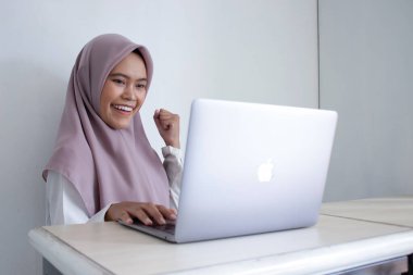 Mor başörtüsü takan Asyalı genç bir kadın gülümsüyor ve dizüstü bilgisayara bakarken ellerini havada tutuyor.