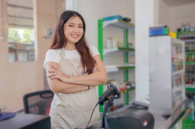 Gülümseyen Asyalı genç kadın market personeli kollarını kavuşturup vitrin rafının önünde duruyor.