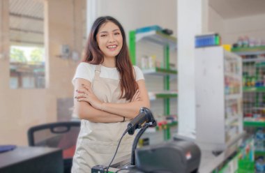 Gülümseyen Asyalı genç kadın market personeli kollarını kavuşturup vitrin rafının önünde duruyor.