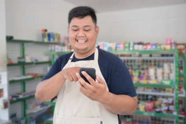 Market çalışanı Asyalı erkek çalışanı gülümseyerek onaylanmak için el kol hareketi yapıyor ve gri önlükle vitrin rafının önünde dikilirken telefona bakıyor.