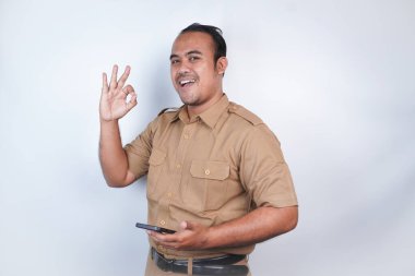 Asyalı bir devlet memuru ya da devlet memuru kahverengi üniformalı bir jest yapar, telefonunu tutar ve poz verir. Beyaz arka planda. Endonezya PNS, ASN