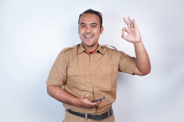 Asyalı bir devlet memuru ya da devlet memuru kahverengi üniformalı bir jest yapar, telefonunu tutar ve poz verir. Beyaz arka planda. Endonezya PNS, ASN