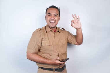 Asyalı bir devlet memuru ya da devlet memuru kahverengi üniformalı bir jest yapar, telefonunu tutar ve poz verir. Beyaz arka planda. Endonezya PNS, ASN