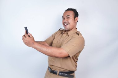 Asyalı devlet memurları veya devlet memurları gülümseyen bir adam kahverengi üniformalı bir jestle telefonunu gösterir. Beyaz arka planda. Endonezya PNS, ASN