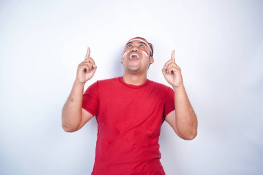 Kırmızı tişört giyen, bayrak bandı takan, yanındaki fotokopi alanını gösteren, beyaz arka planla izole edilmiş, şok olmuş Asyalı bir adam. Endonezya 'nın bağımsızlık günü konsepti.