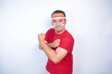 Kollarını ve kaslarını kaldırarak güçlü bir hareket gösteren genç bir Asyalı adam kırmızı gömlek ve beyaz arka planla izole edilmiş bantla gururla gülümsüyor. Endonezya 'nın bağımsızlık günü konsepti.