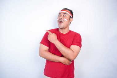 Kırmızı tişört giyen, bayrak bandı takan, yanındaki fotokopi alanını gösteren, beyaz arka planla izole edilmiş, şok olmuş Asyalı bir adam. Endonezya 'nın bağımsızlık günü konsepti.