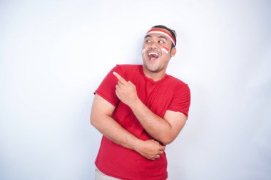 Kırmızı tişört giyen, bayrak bandı takan, yanındaki fotokopi alanını gösteren, beyaz arka planla izole edilmiş, şok olmuş Asyalı bir adam. Endonezya 'nın bağımsızlık günü konsepti.