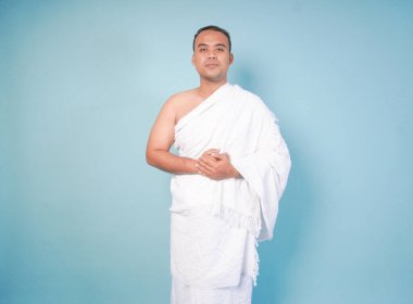 İhram giyen genç Asyalı Müslüman adam izole edilmiş mavi arka plan pozu veriyor. Hac konsepti. İslam