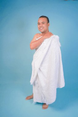 İhram takan Asyalı Müslüman genç adam, ihramın nasıl kullanılacağını izole edilmiş mavi arka planda gösteriyor. Hac konsepti. İslam Müslüman duruşu