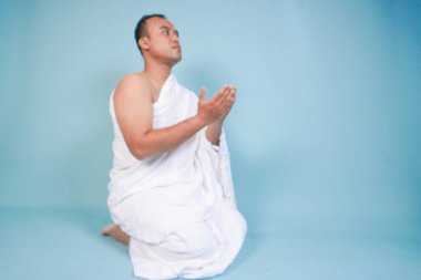 İhramda oturan ve el ele tutuşup dua eden ciddi bir Asyalı müslüman. Mavi arka planda izole edilmiş el hareketleriyle. Hac konsepti. İslam Müslüman konsepti