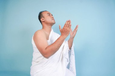 İhramda oturan ve el ele tutuşup dua eden ciddi bir Asyalı müslüman. Mavi arka planda izole edilmiş el hareketleriyle. Hac konsepti. İslam Müslüman konsepti