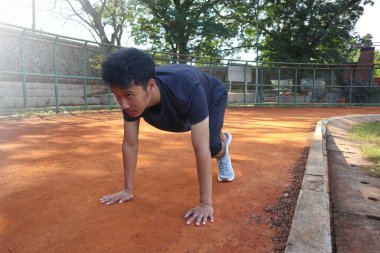 Genç Asyalı koşucu toprak sahada başlangıç pozisyonunda koşmaya hazır. Adam gri bir gömlek ve siyah bir şort giyiyor.