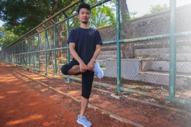 Bir adam kirli bir arazide bacağını esnetiyor. Adam siyah gömlek ve siyah şort giyiyor.