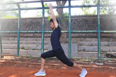 Adamın biri toprak alanda yoga pozu veriyor. Adam siyah bir gömlek ve şort giyiyor.