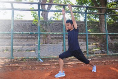 Adamın biri toprak alanda yoga pozu veriyor. Adam siyah bir gömlek ve şort giyiyor.