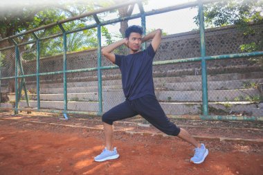 Adamın biri toprak alanda yoga pozu veriyor. Adam siyah bir gömlek ve şort giyiyor.