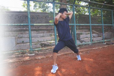 Adamın biri toprak alanda yoga pozu veriyor. Adam siyah bir gömlek ve şort giyiyor.