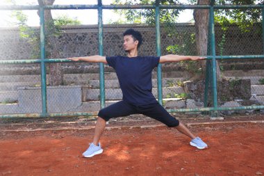 Adamın biri toprak alanda yoga pozu veriyor. Adam siyah bir gömlek ve şort giyiyor.