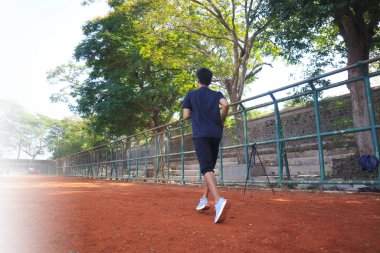 Adamın biri arkasında çitle pis bir arazide koşuyor. Adam siyah gömlek ve siyah şort giyiyor.