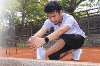 Bir adam kirli bir arazide bacağını esnetiyor. Adam gri bir gömlek ve siyah bir şort giyiyor.