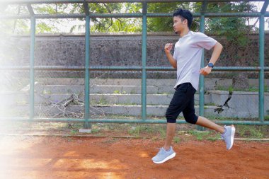 Arkasında yeşil bir çitle toprak bir alanda koşan bir adam var. Adam gri bir gömlek ve siyah bir şort giyiyor.