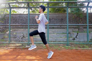 Arkasında yeşil bir çitle toprak bir alanda koşan bir adam var. Adam gri bir gömlek ve siyah bir şort giyiyor.