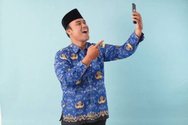 Kopri Blue Batik üniforması giyen gülümseyen devlet çalışanı akıllı telefonunu işaret ederken görüntülü arama yapıyor. PNS, ASN memur konsepti.