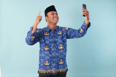 Kopri Blue Batik üniforması giyen gülümseyen devlet çalışanı akıllı telefonunu işaret ederken görüntülü arama yapıyor. PNS, ASN memur konsepti.