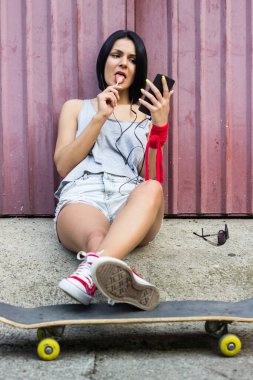 Hipster girl sitting on her skateboard and using smartphone