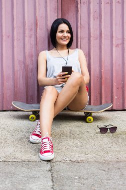 Hipster girl sitting on her skateboard and using smartphone