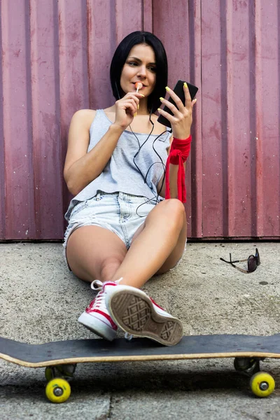 Hipster girl sitting on her skateboard and using smartphone