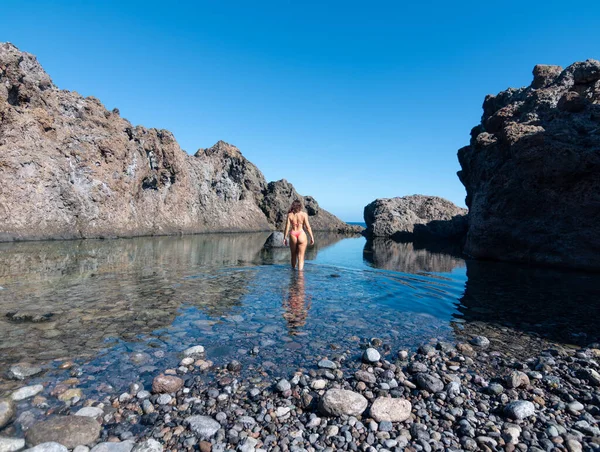 Young woman from behind in a bikini entering the water of a natural pool of volcanic rock in Tenerife