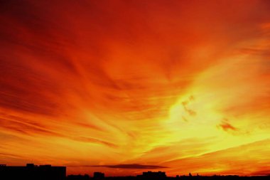 Canlı bir günbatımı, gökyüzünü turuncu ve sarıya boyar. Bulutlar siluetli bir şehrin ufuk çizgisinin üzerinde karmaşık desenler oluşturur..
